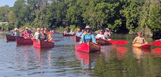 A variety of people in red canoes on a river with green trees behind them.