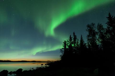 photo of the northern lights over a lake with evergreen trees on the side