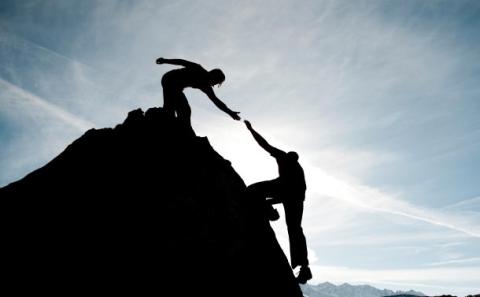 Photo in silhouette of one person reaching to help another person get to the top of a summit