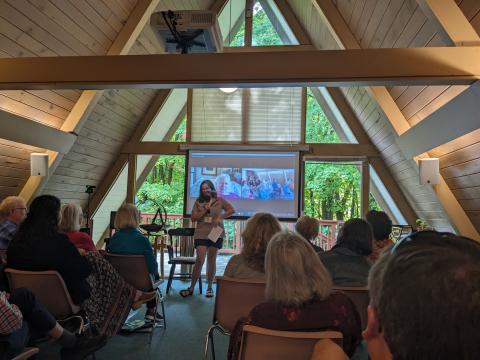 A person presenting in front of a screen, to a group of seated people inside an a-frame building.