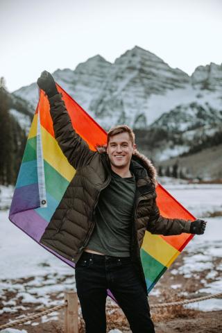 Mikah Meyer standing in front of a mountain, holding a pride flag and smiling