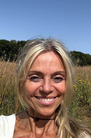 Head and shouldrs of woman with blonde hair and a smile. The background is a field of golden wheat with green trees behind it and a clear blue sky.