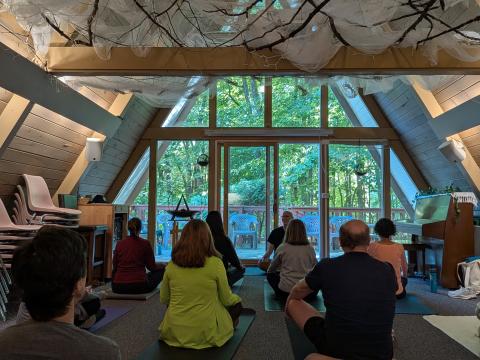 A group of people sit on yoga mats on the floor of an A-frame room, facing a bald man with glasses seated in front of tall windows, trees visible through the glass and gauzy fabric and bare branches hung across the ceiling.